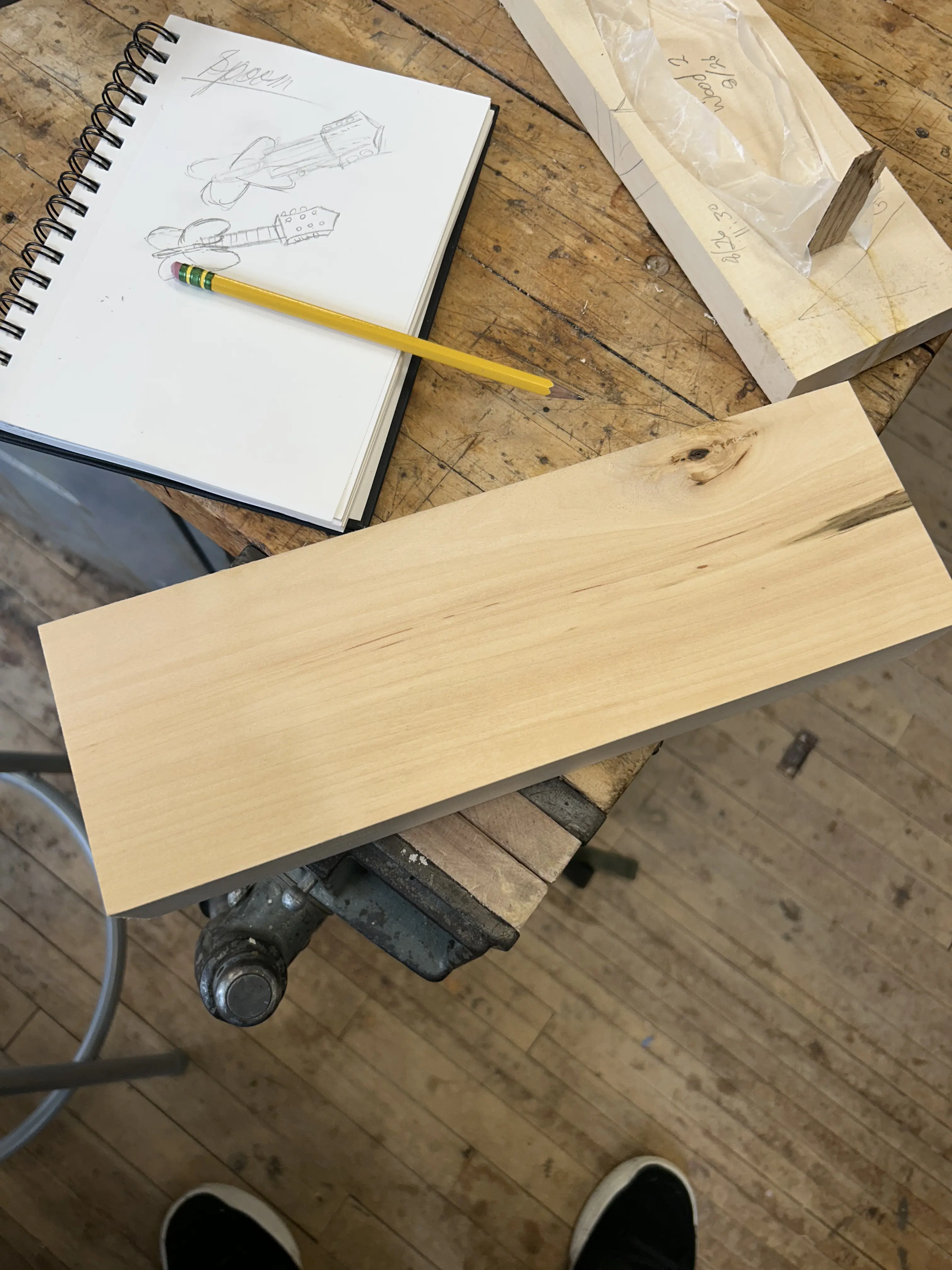 A piece of wood following the jointing and planing processes sitting on a table next to a sketchbook showcasing a drawing of a guitar with a butterfly shaped body.